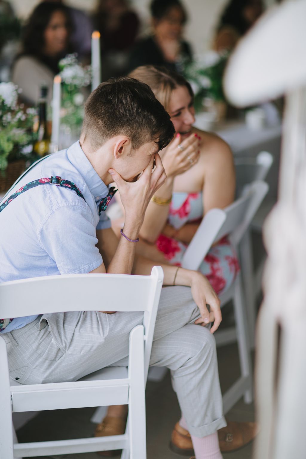 Groom in light blue shirt bows head in prayer during ceremony, with bride and guests visible behind him