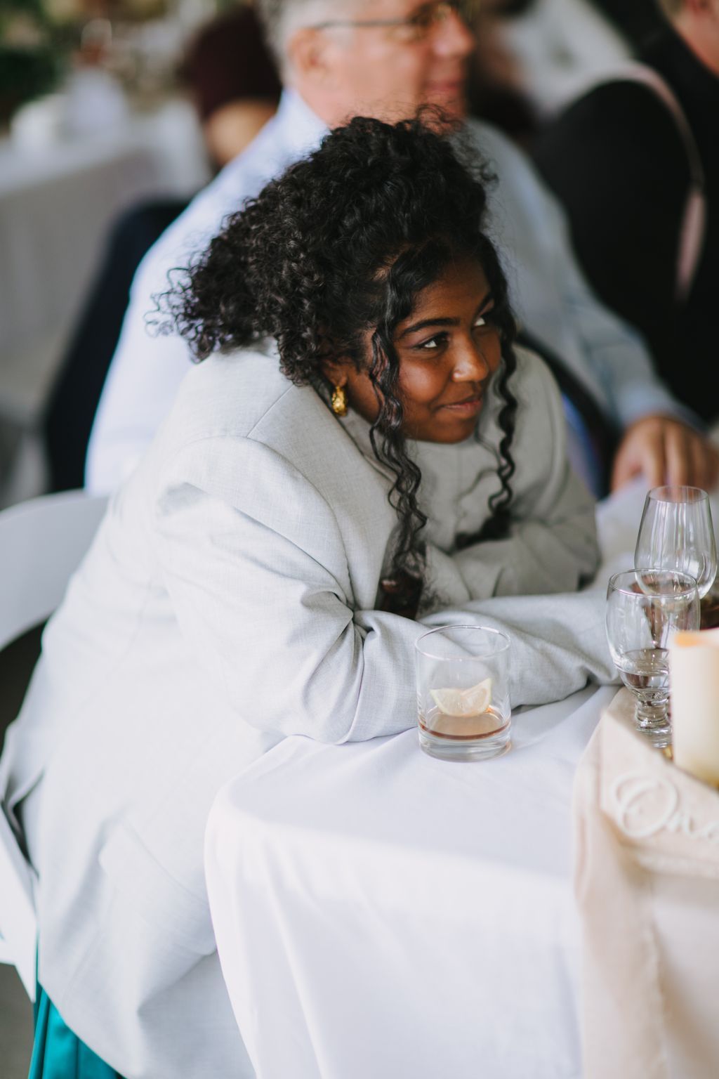Woman in white blazer with curled hair smiles over shoulder at formal event, holding glass of whiskey