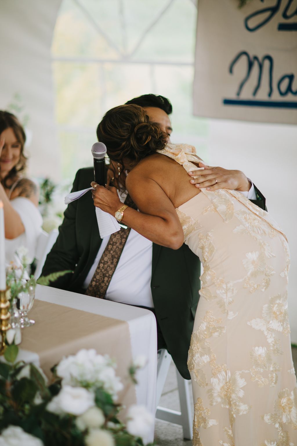 Groom in dark suit holding microphone embraces bride in beaded gown during wedding ceremony, soft natural light from windows behind them