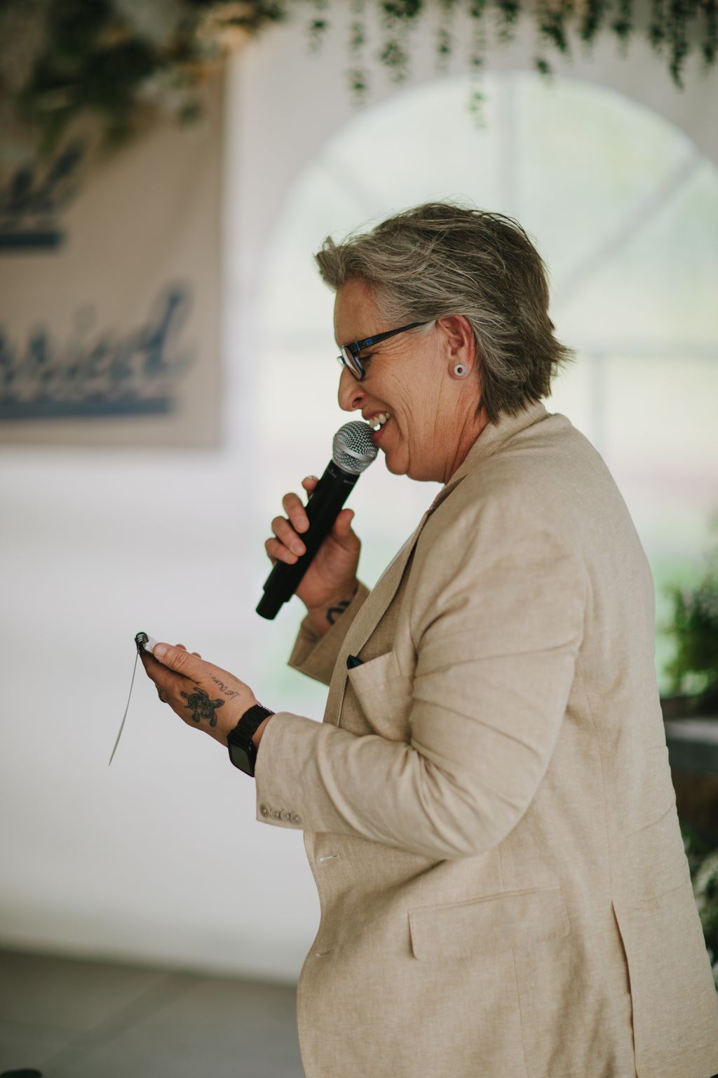 Woman in tan blazer speaking into microphone at wedding reception, smiling warmly