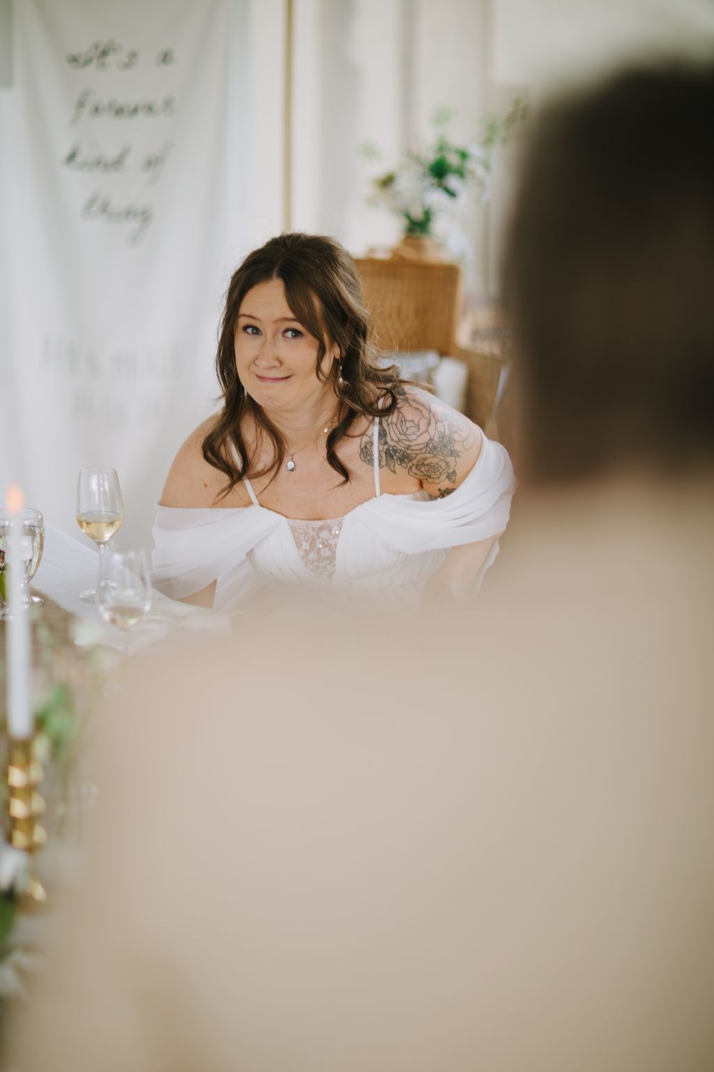 Bride with dark wavy hair and shoulder tattoo smiles softly in off-shoulder white lace gown in bright, minimalist interior
