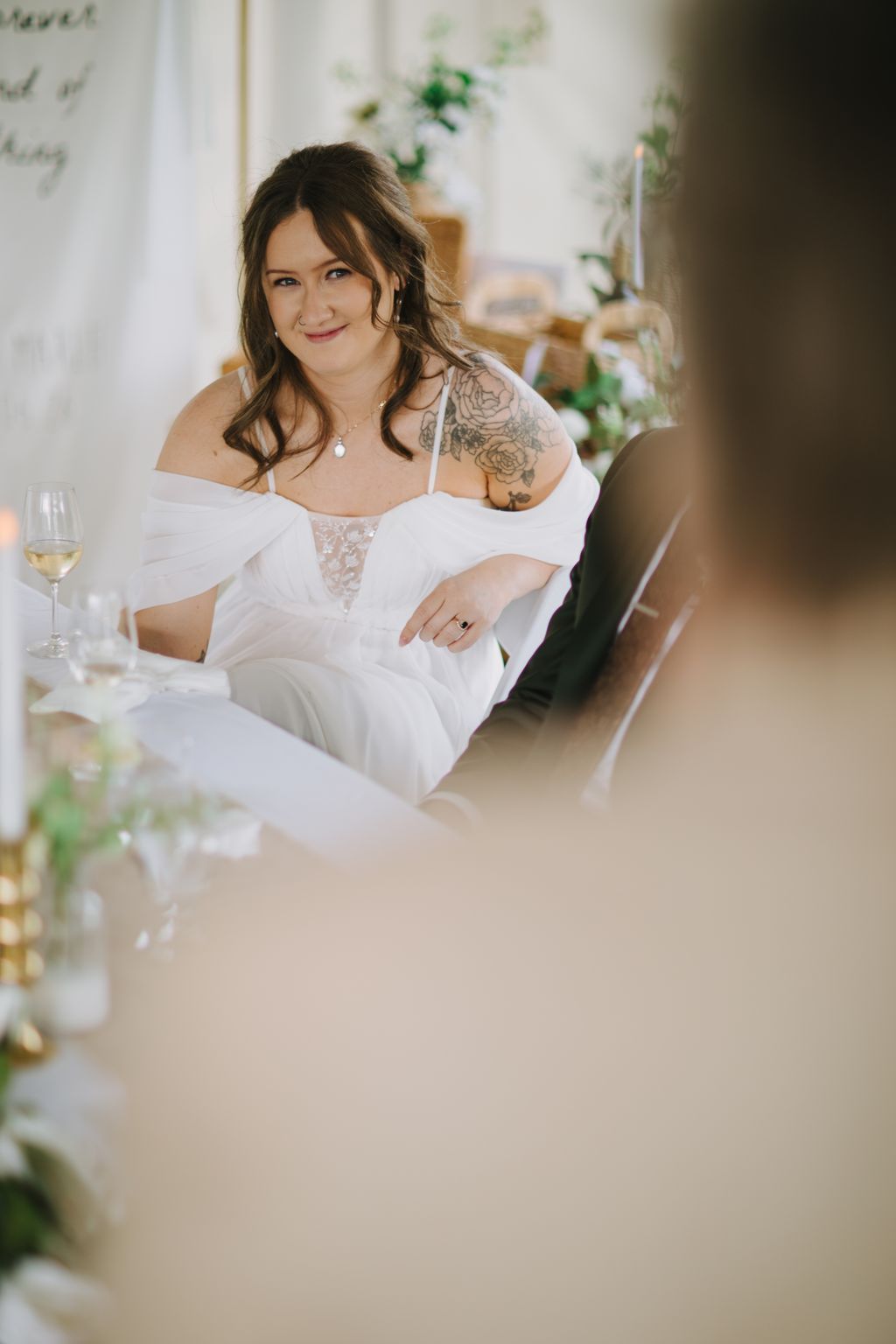 Bride in white off-shoulder lace dress with rose tattoo smiles at camera in bright, plant-filled indoor setting