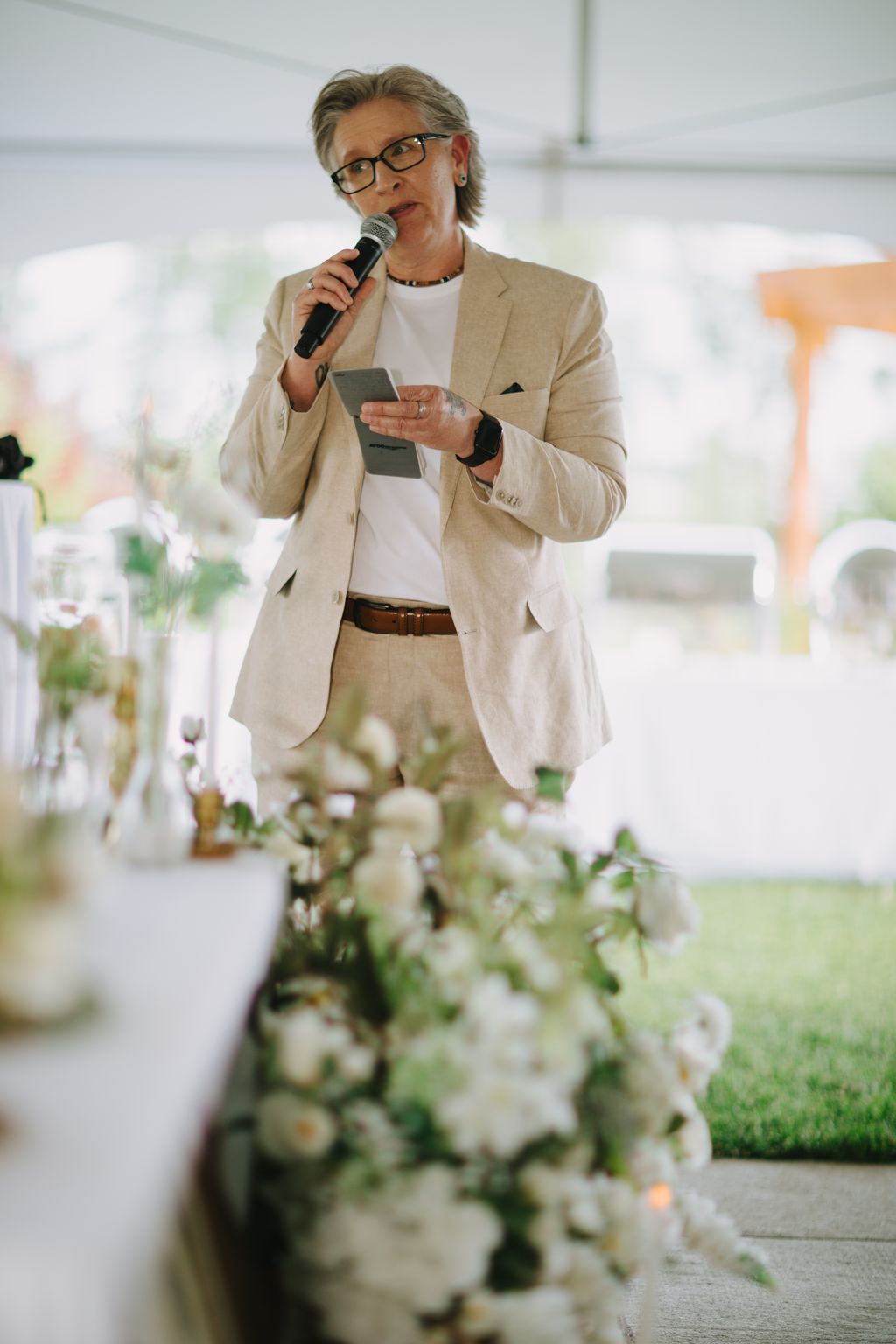 Woman in beige blazer speaking at microphone during wedding reception with white flowers in foreground