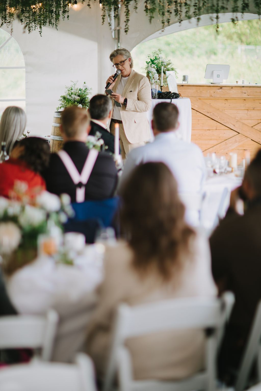 Woman in beige blazer speaking at microphone during elegant indoor wedding reception with hanging greenery and floral arrangements