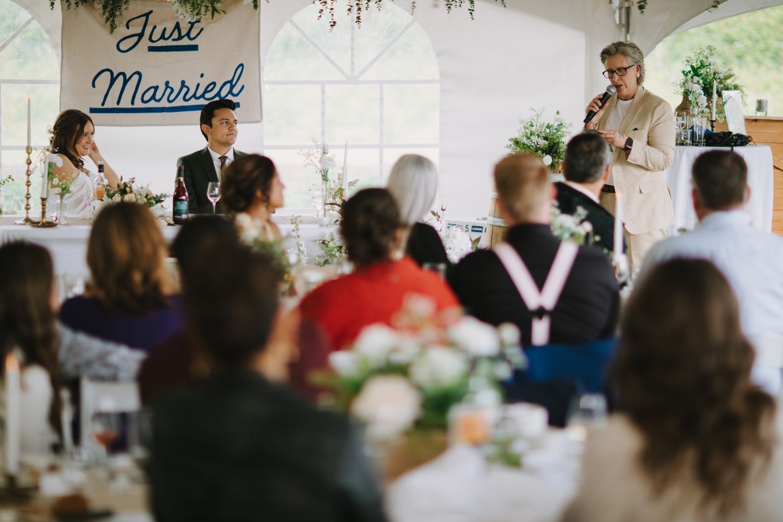 Guests seated at a reception table with fresh flowers, sunlight streaming through arched windows behind them