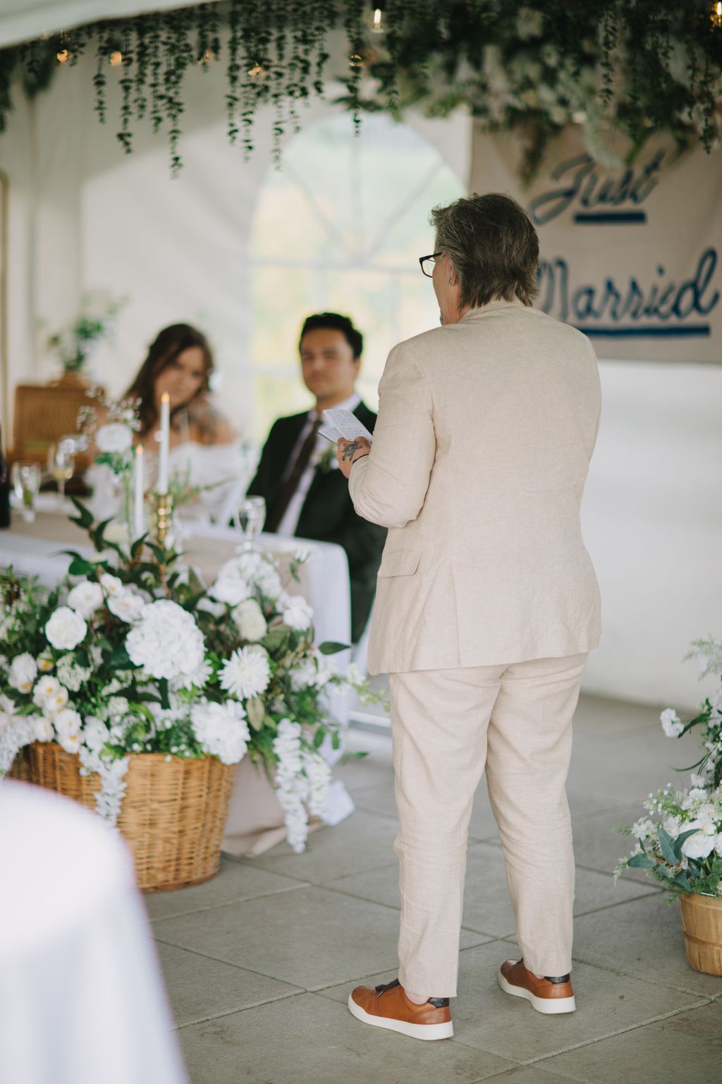 Man in cream suit speaking at wedding ceremony with hanging greenery and white floral arrangements overhead