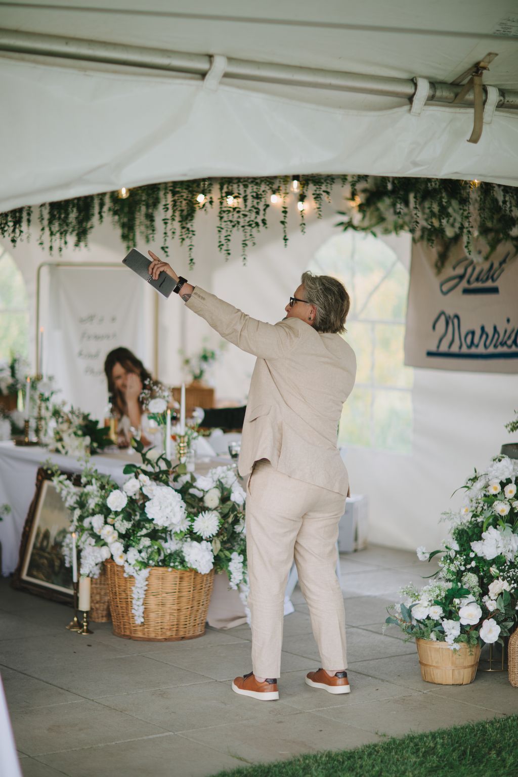 Man in beige suit toasting under white tent with hanging greenery and string lights during outdoor wedding reception