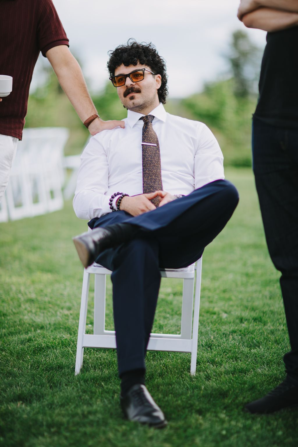 Groom in white shirt and patterned tie sits relaxed on white chair in grass, wearing sunglasses and purple beaded bracelets