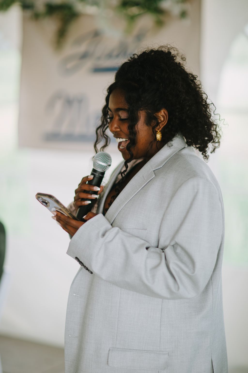 Person in white blazer holding microphone, smiling with eyes closed, golden sunlight, blurred outdoor background