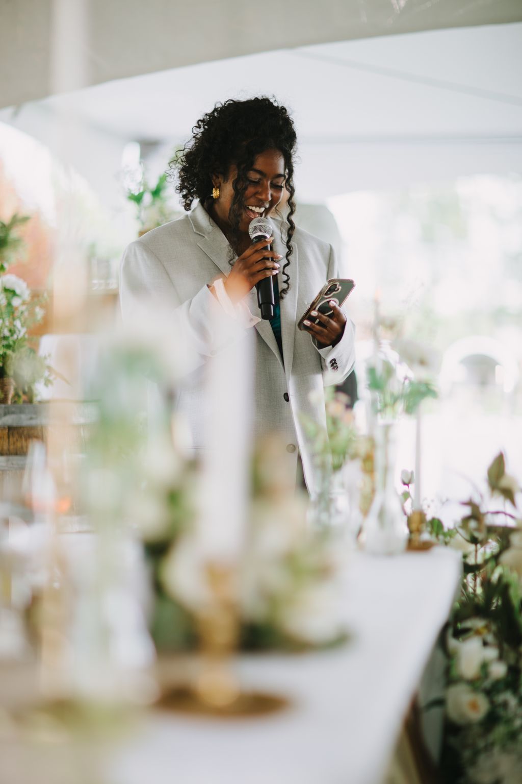 Bride in cream suit holding microphone and phone, smiling during wedding ceremony with blurred floral arrangements in foreground