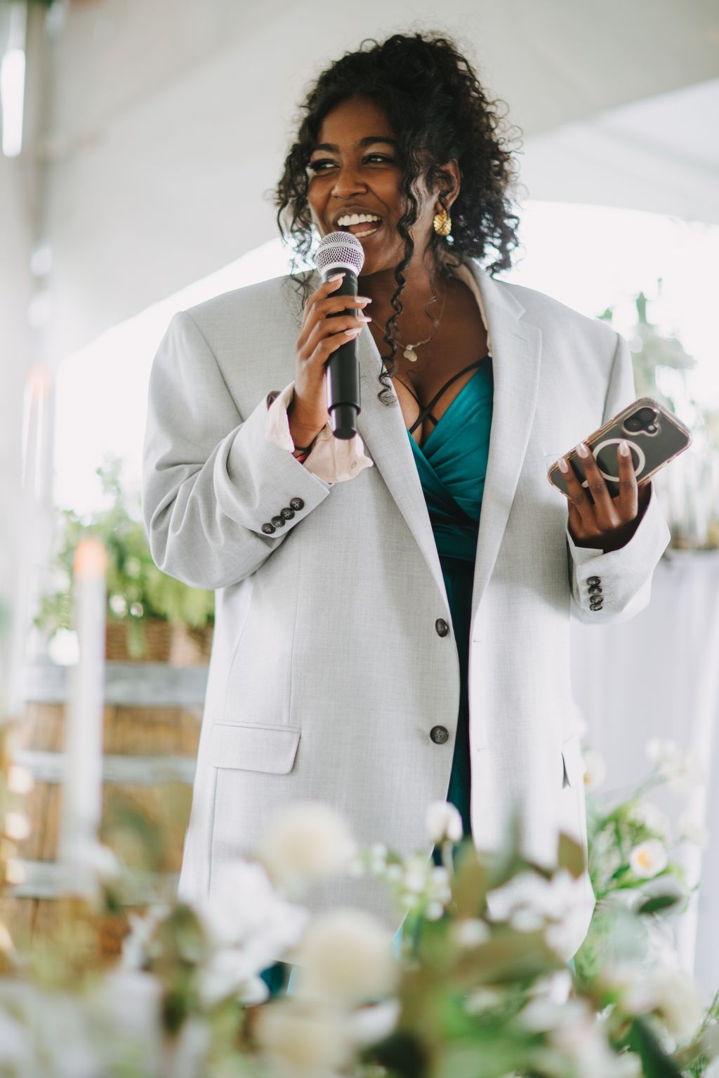 Woman in cream blazer and teal top speaking into microphone at outdoor wedding ceremony, smiling brightly with soft natural light