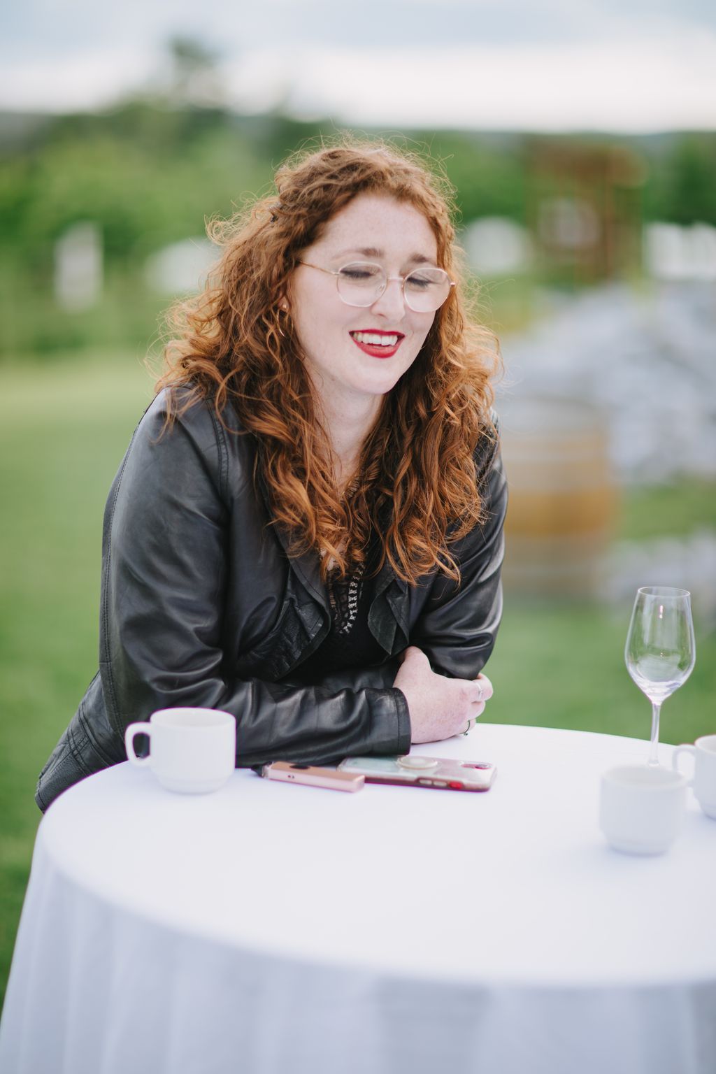 Woman with curly red hair and glasses smiles outdoors at white table with blurred green background