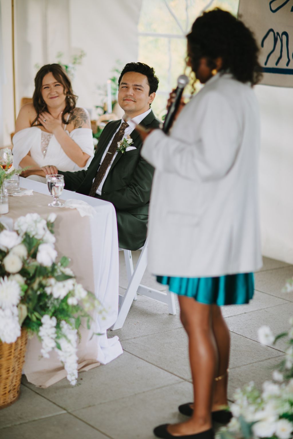 Groom in dark suit with boutonniere gazes ahead during wedding ceremony in bright, plant-decorated venue
