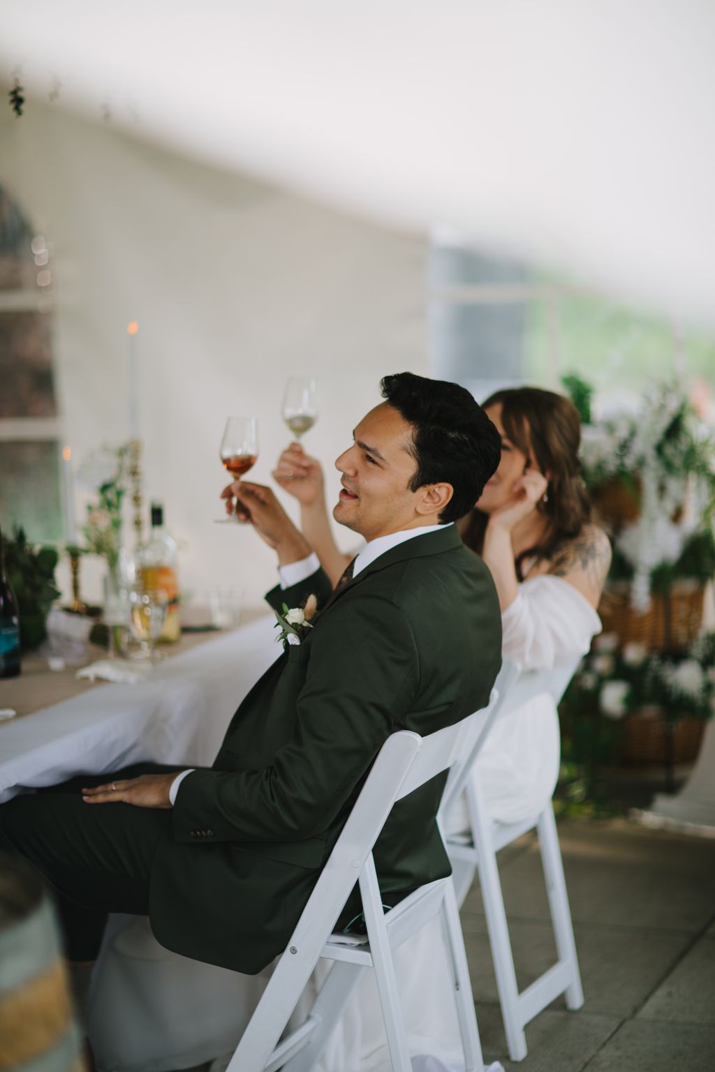 Groom in dark suit raising wine glass during reception toast, seated beside bride in white dress, bright indoor venue