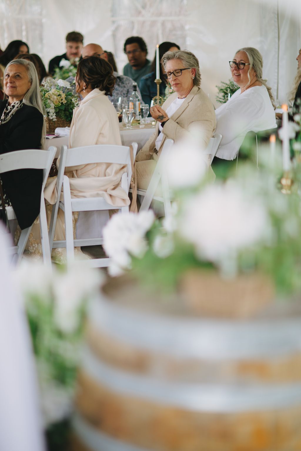 Woman in beige blazer and glasses seated at elegant outdoor wedding reception with white flowers and place settings in soft focus foreground