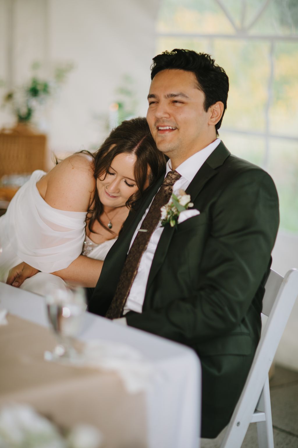 Groom in dark suit with patterned tie smiles while bride leans against him in white dress, soft natural light in blurred indoor setting