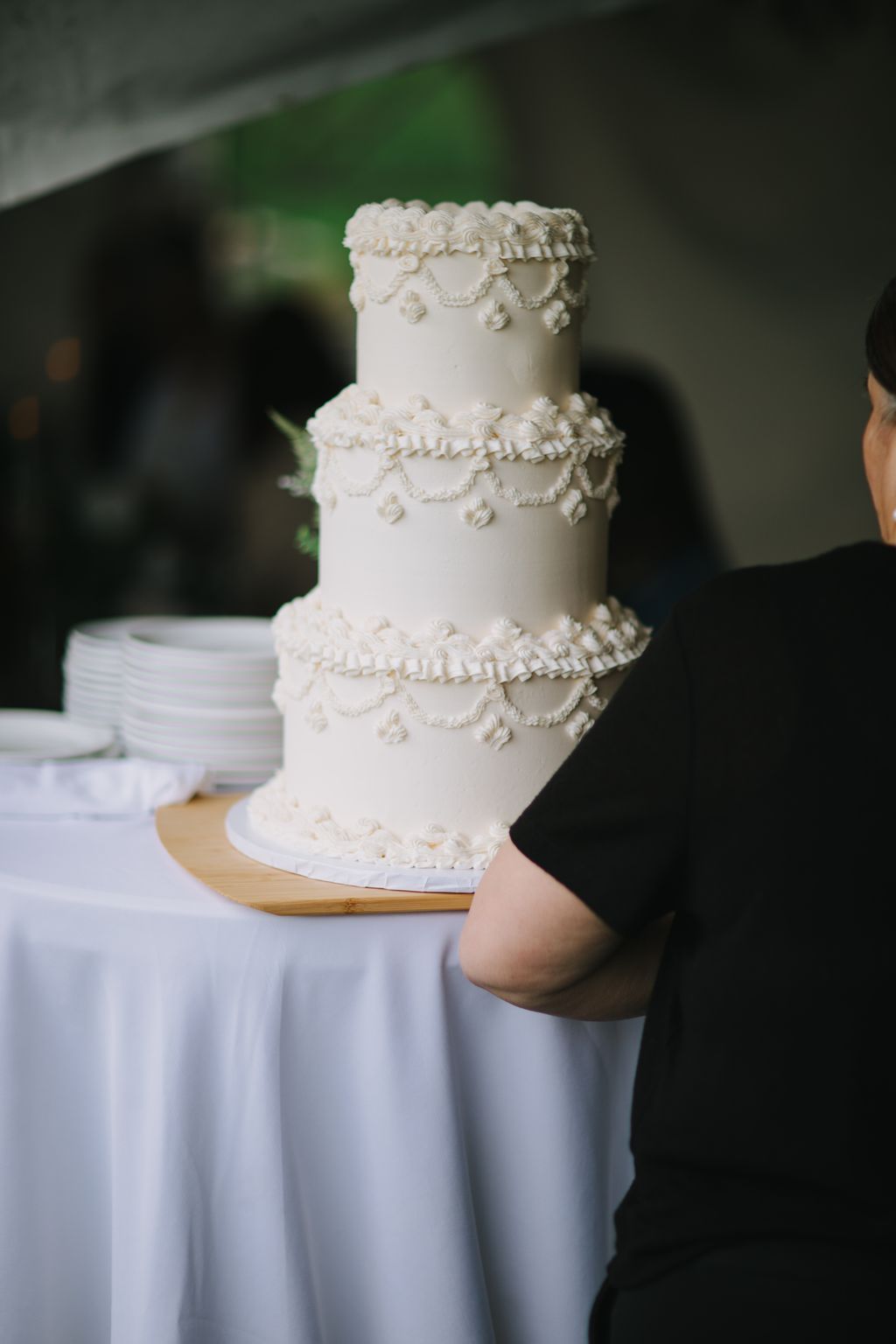 Three-tiered ivory wedding cake with piped ruffle borders and pearl swags, held by person in light blue dress