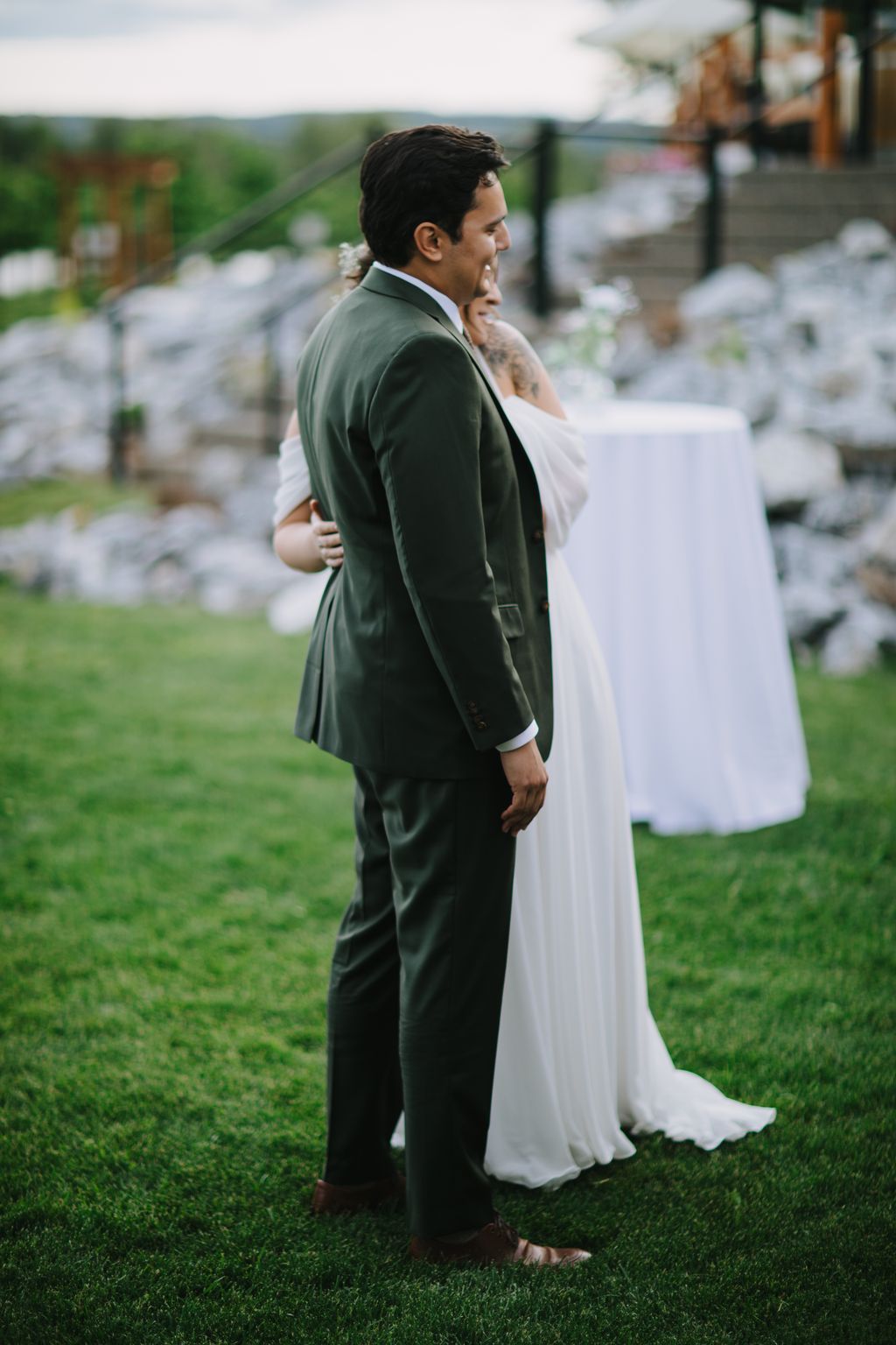 Groom in sage green suit embraces bride in white dress on grass with blurred garden backdrop