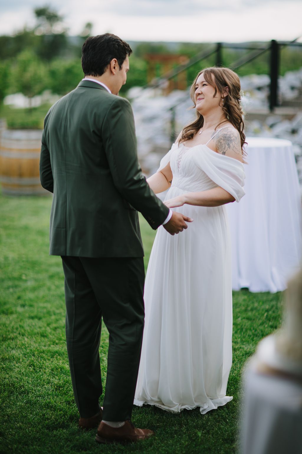 Pregnant bride in flowing white dress and groom in olive suit hold hands outdoors with blurred garden in background
