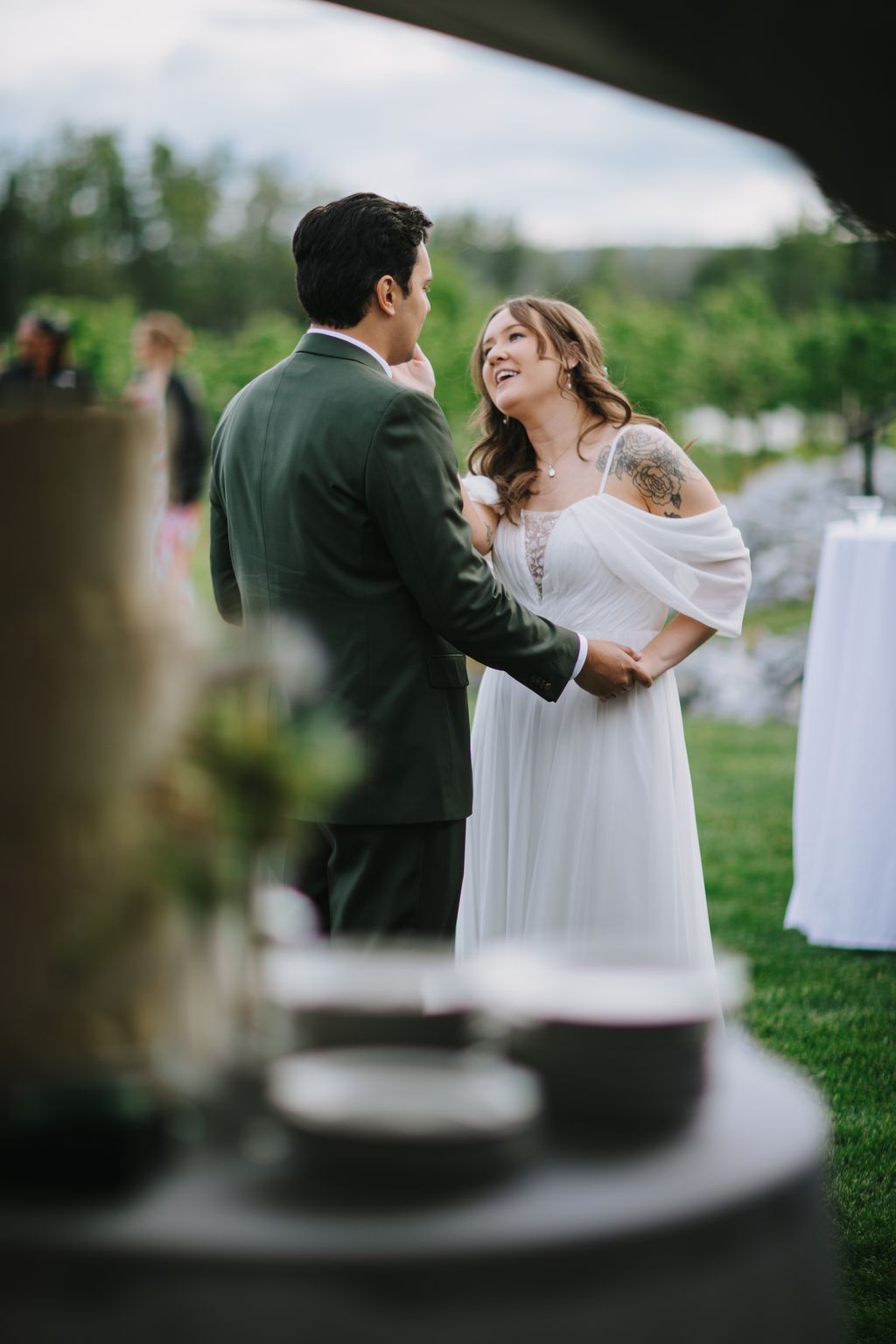 Bride in white off-shoulder dress and groom in sage green suit face each other smiling outdoors with green landscape and covered structure behind them