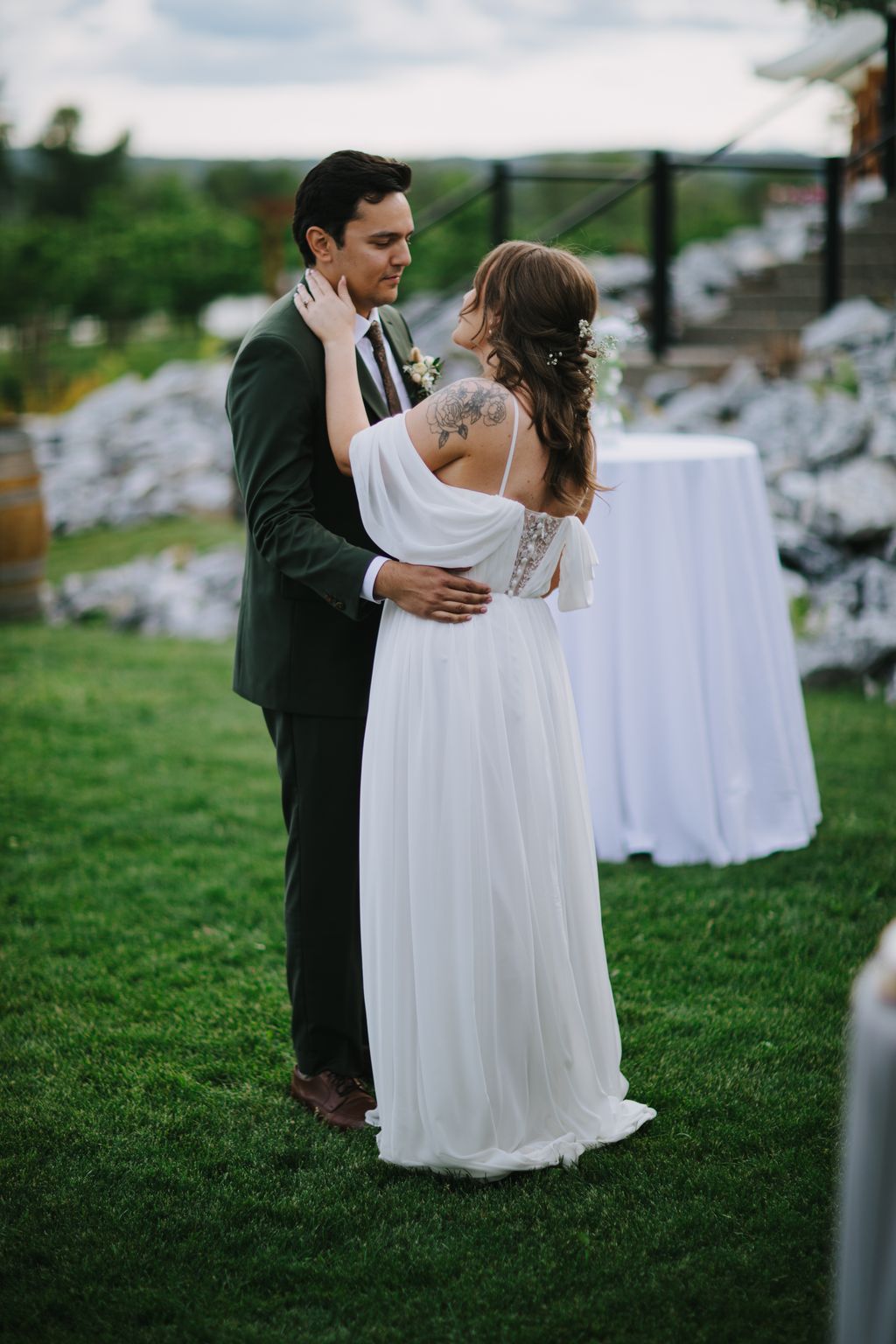 Groom in olive suit embracing bride in flowing white dress on manicured lawn with pastoral backdrop