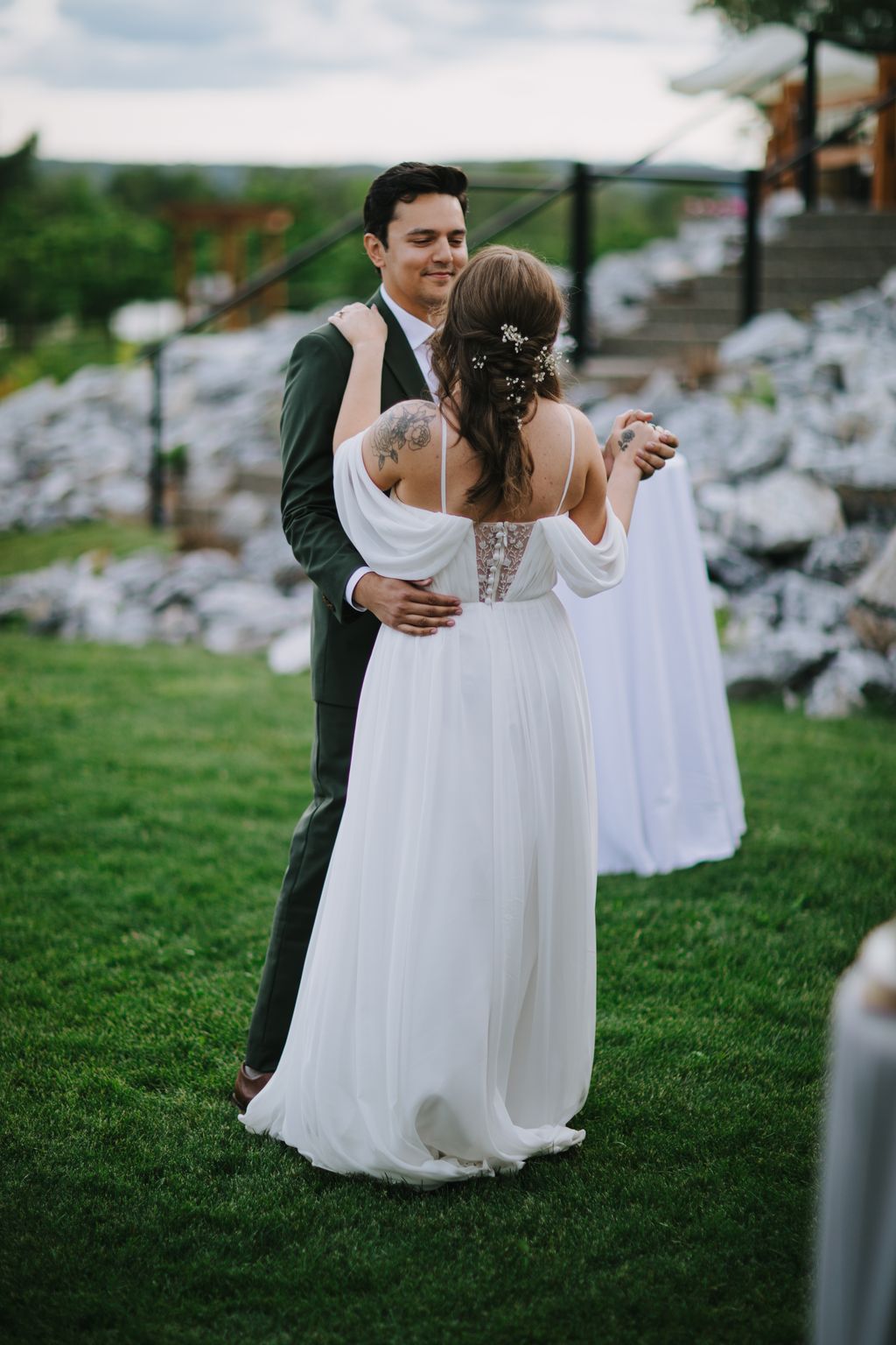 Groom in sage green suit embraces bride in flowing white dress with floral hair pins on grassy outdoor venue at golden hour