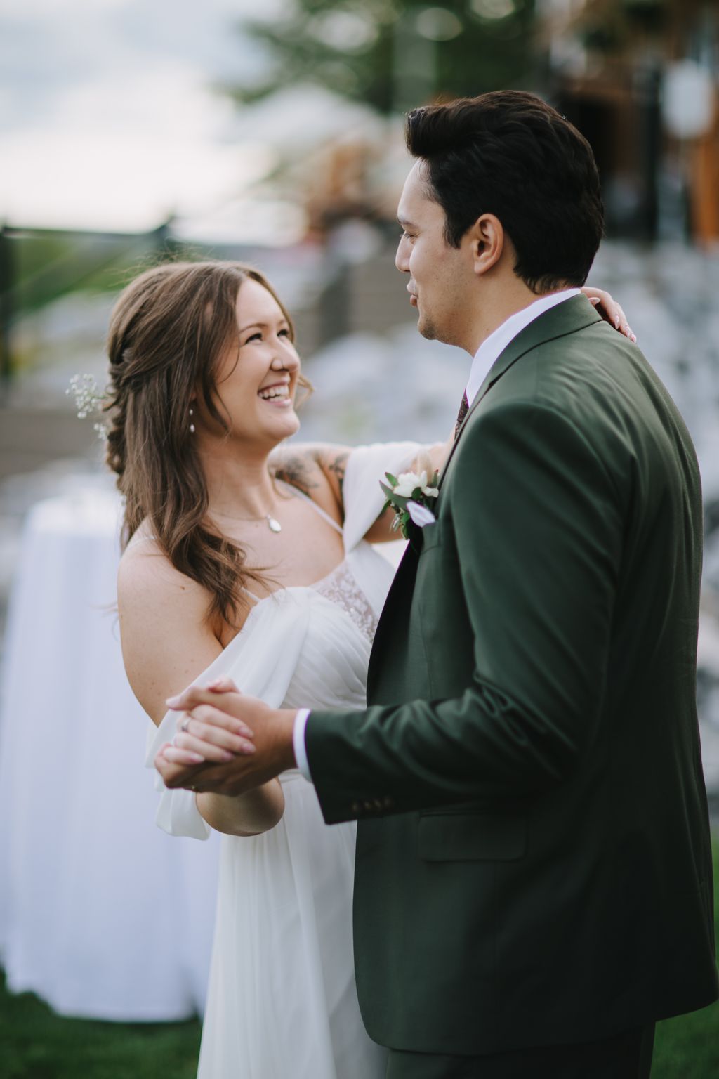 Bride in white dress and groom in green suit smile at each other during outdoor ceremony with soft natural light