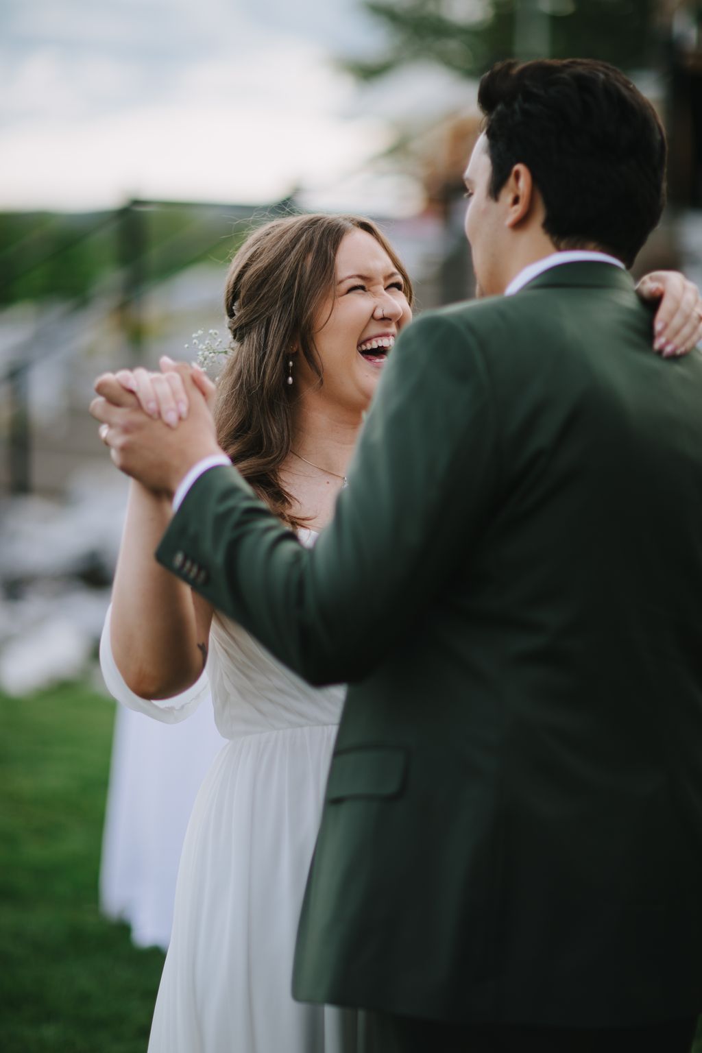 Bride in white dress laughing with groom in green suit during outdoor ceremony, natural light