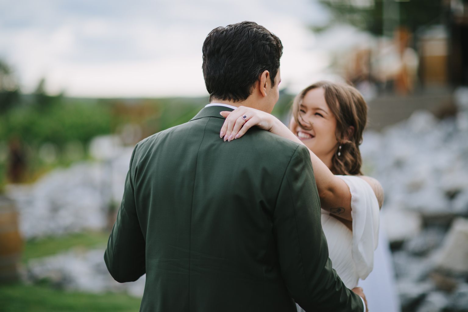 Bride''s hand with wedding ring rests on groom''s shoulder from behind, green field backdrop, soft natural light