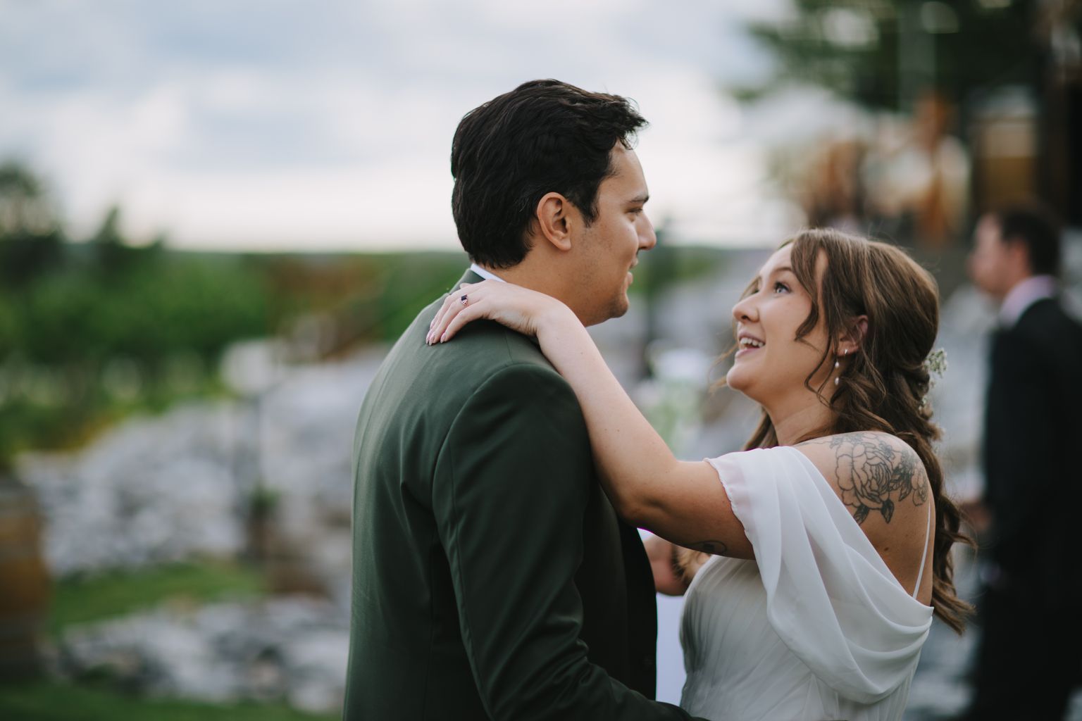 Groom in olive shirt embraces bride from behind in outdoor setting with soft overcast light