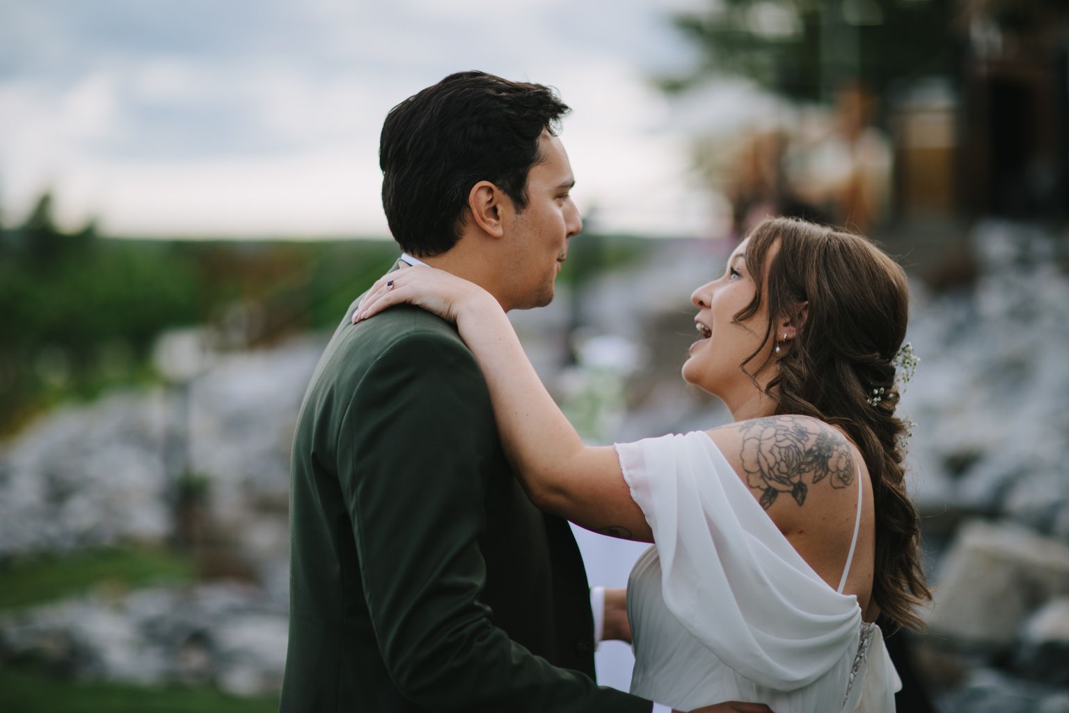 Groom in olive green shirt gazes toward bride in white, her hand on his shoulder, soft outdoor lighting