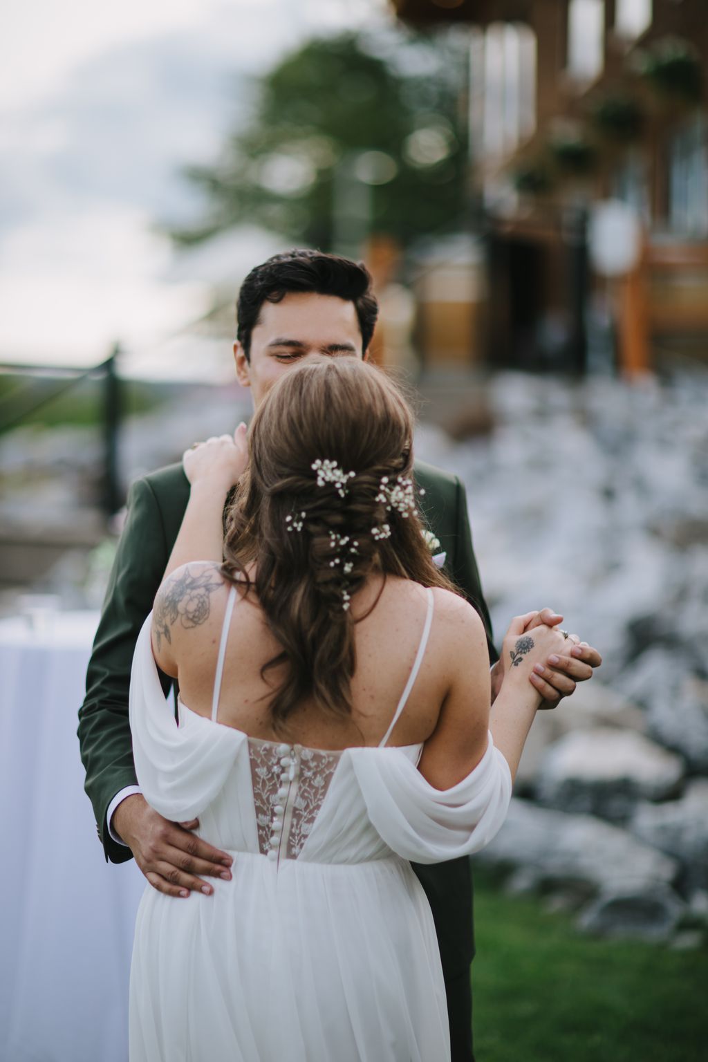 Bride in white dress with baby''s breath hair flowers seen from behind, groom in green jacket beside her in soft outdoor light