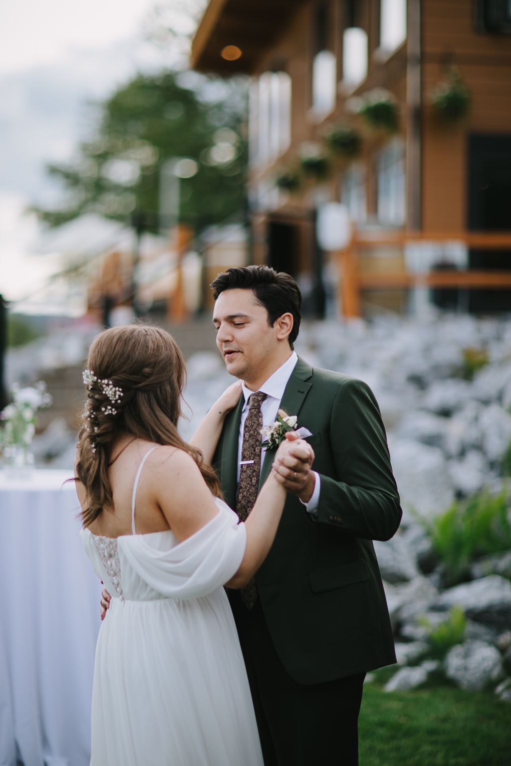 Groom in forest green suit gazes at bride in white dress during outdoor ceremony at elegant venue with hanging lanterns