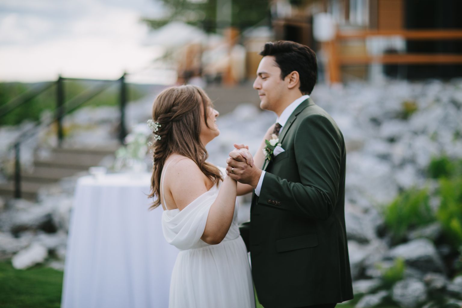 Bride in white draped gown and groom in sage green suit hold hands during outdoor ceremony with soft natural light