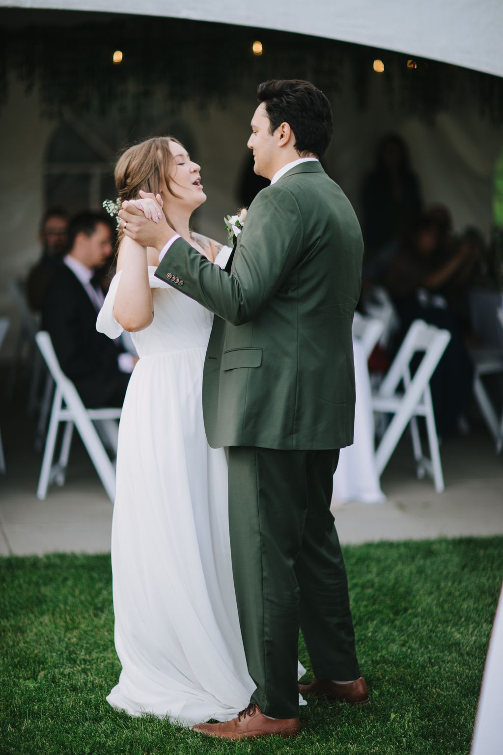 Bride and groom embrace outdoors at dusk, groom in sage green suit, bride in flowing white gown, golden lights glowing softly in background