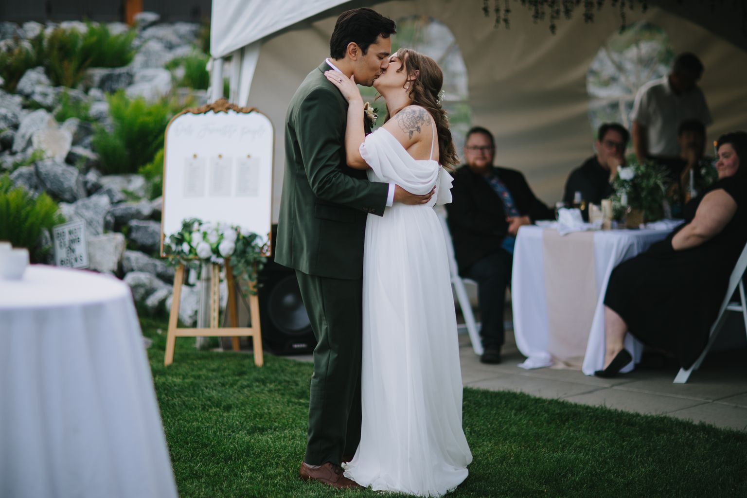 Bride in flowing white gown and groom in sage green suit kiss under a tent at outdoor wedding ceremony