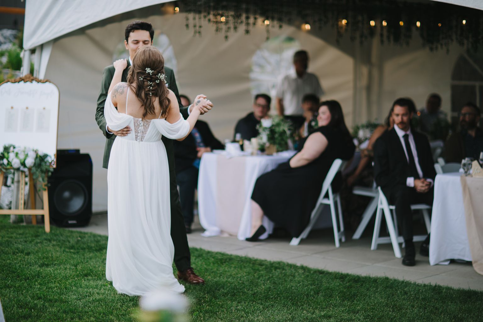 Bride in white dress dancing outdoors at evening reception with guests seated at table under ambient lighting