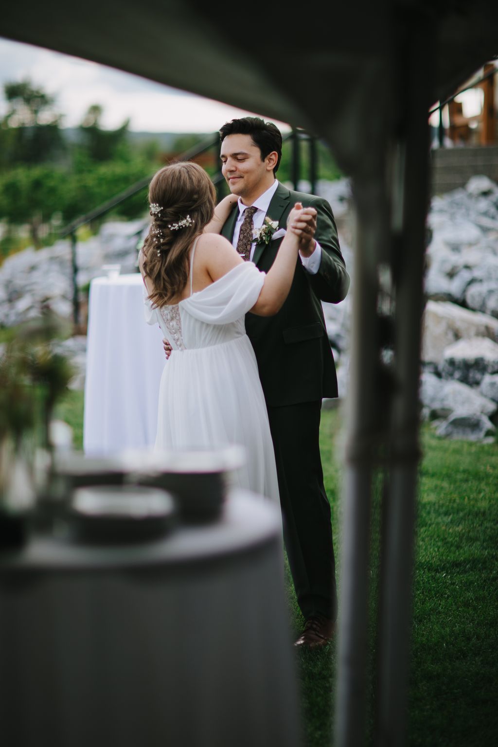 Groom in olive suit dancing with bride in white flowing dress under covered pavilion, soft natural light, intimate moment