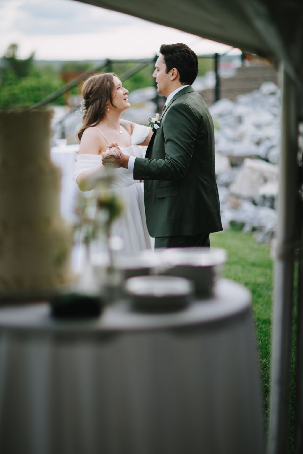 Bride and groom face each other under a green tent, holding hands and smiling, with soft natural light and blurred greenery in the background