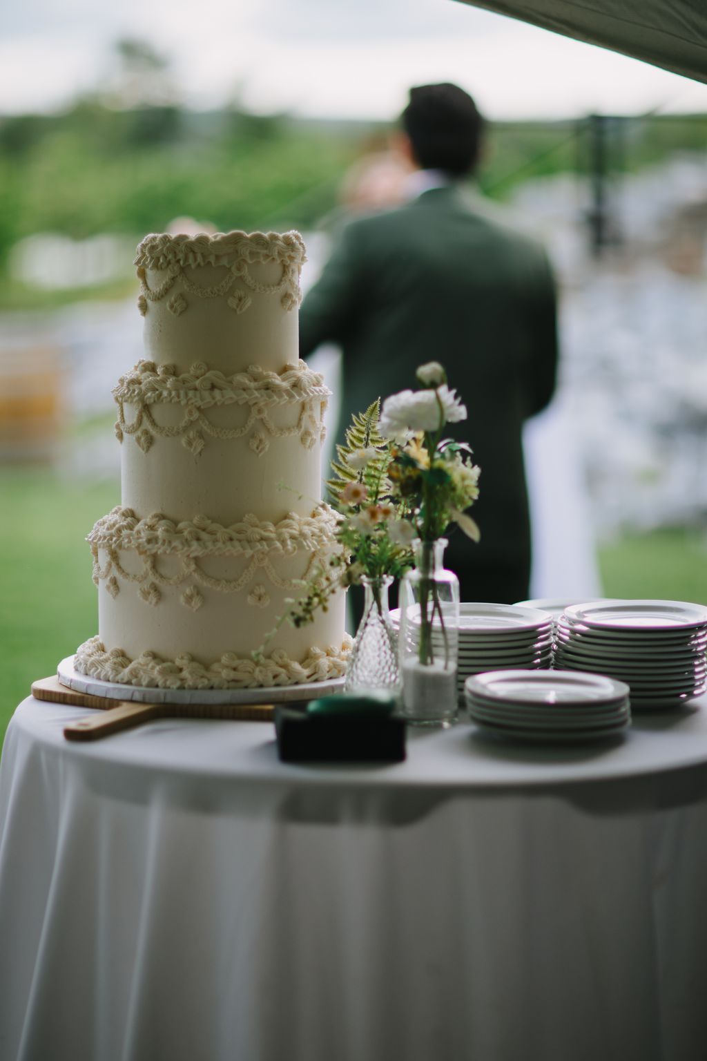 Elegant three-tier wedding cake with intricate piping detail, fresh flowers in glass vases, and blurred couple in background at outdoor reception