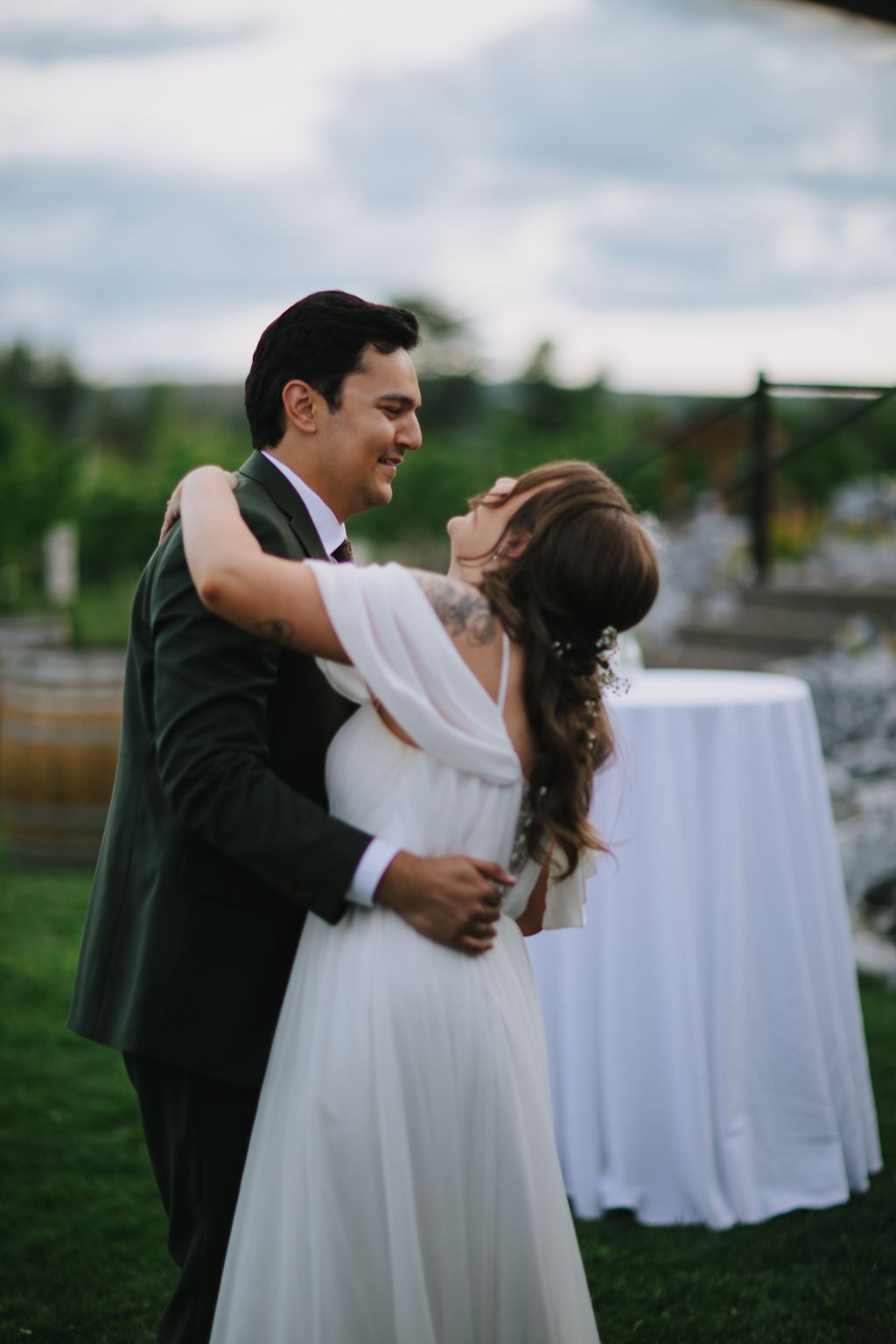 Groom in dark suit and bride in flowing white dress embrace outdoors with soft natural light and blurred green landscape behind them