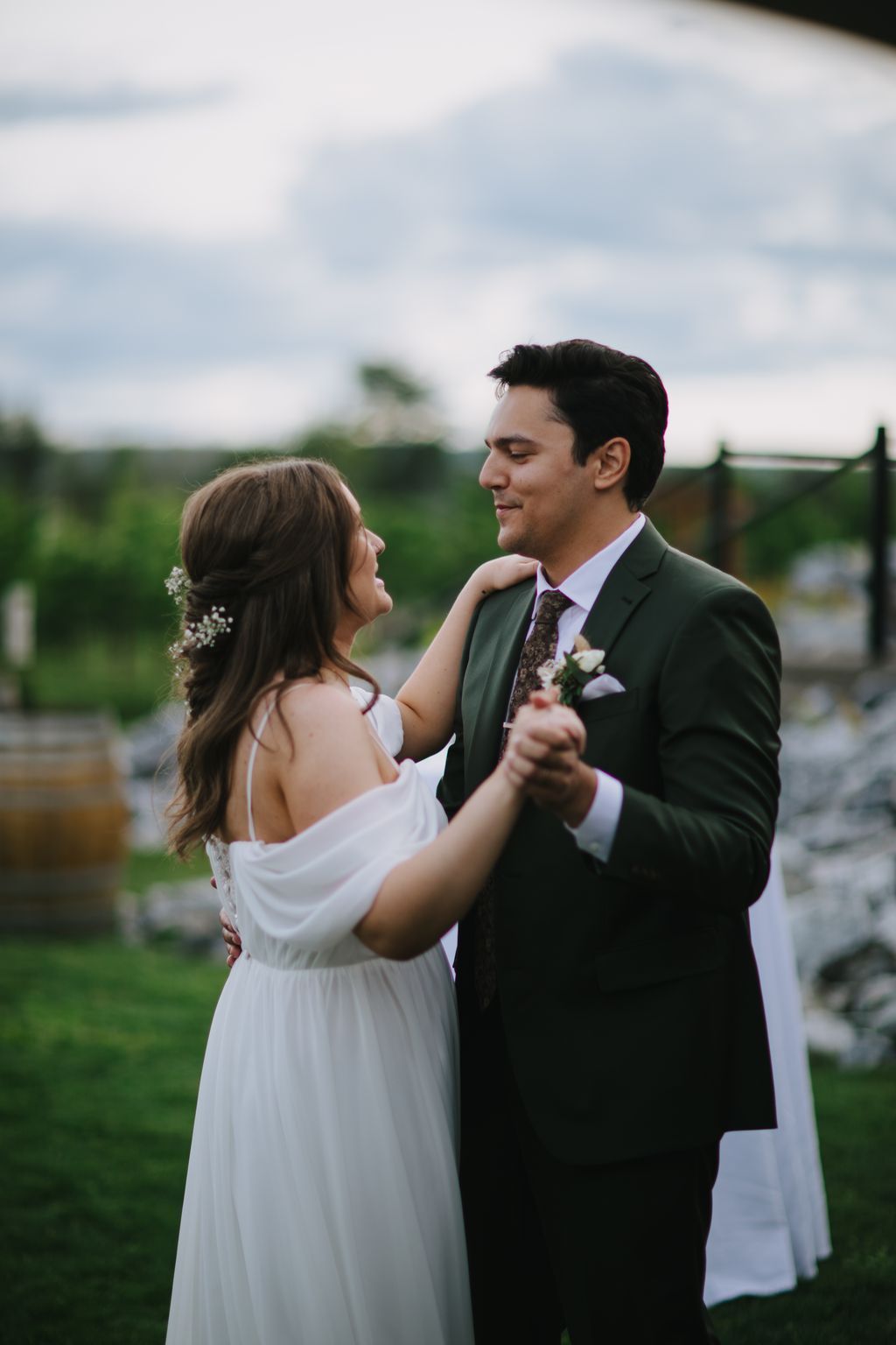 Bride in white off-shoulder dress and groom in sage suit embrace outdoors with blurred green landscape and cloudy sky behind them