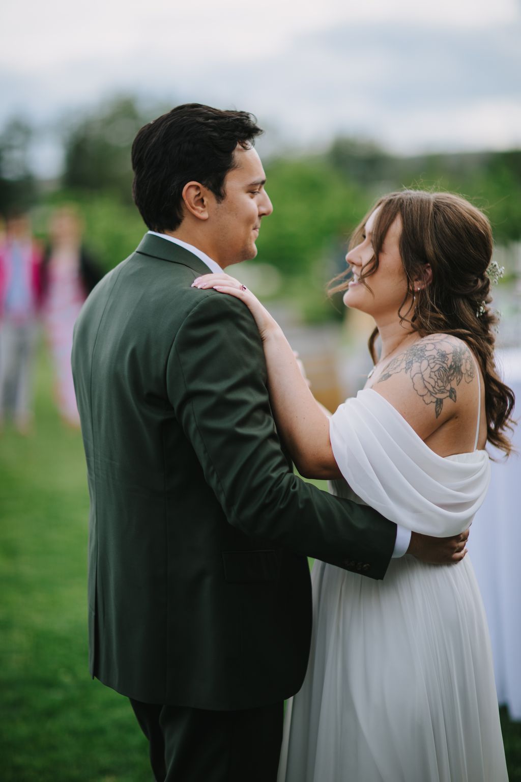 Groom in dark green suit gazes at bride in white dress with hand on his shoulder, soft-focus green field background