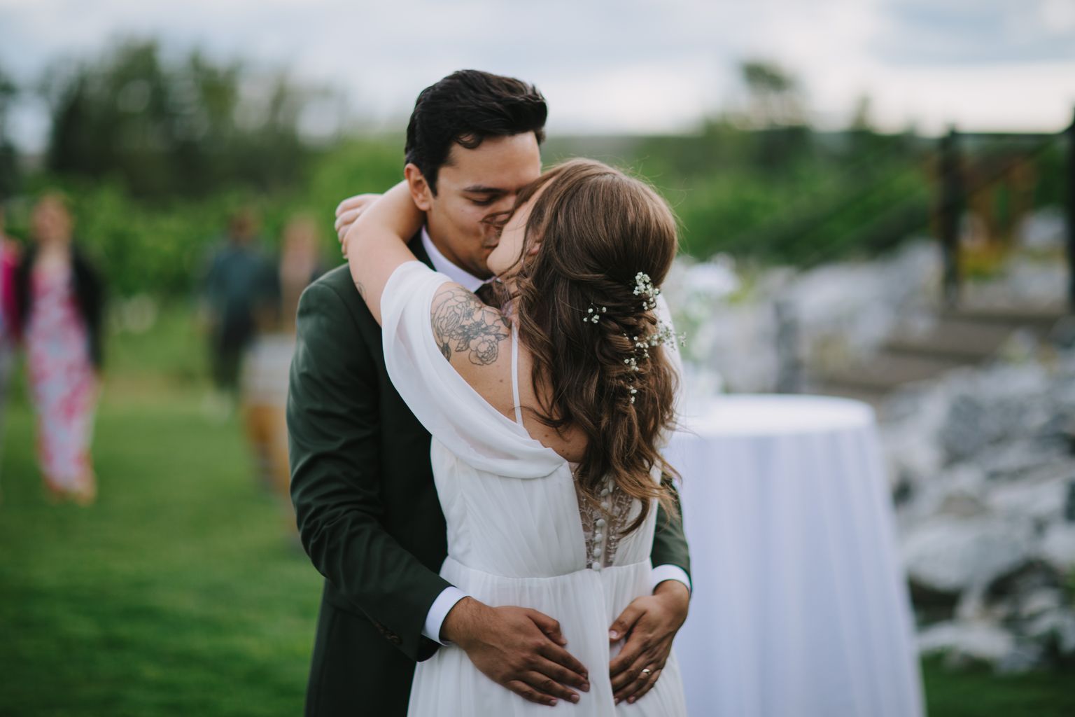 Groom with visible arm tattoo embraces bride in white dress with braided updo adorned with baby''s breath, green field backdrop