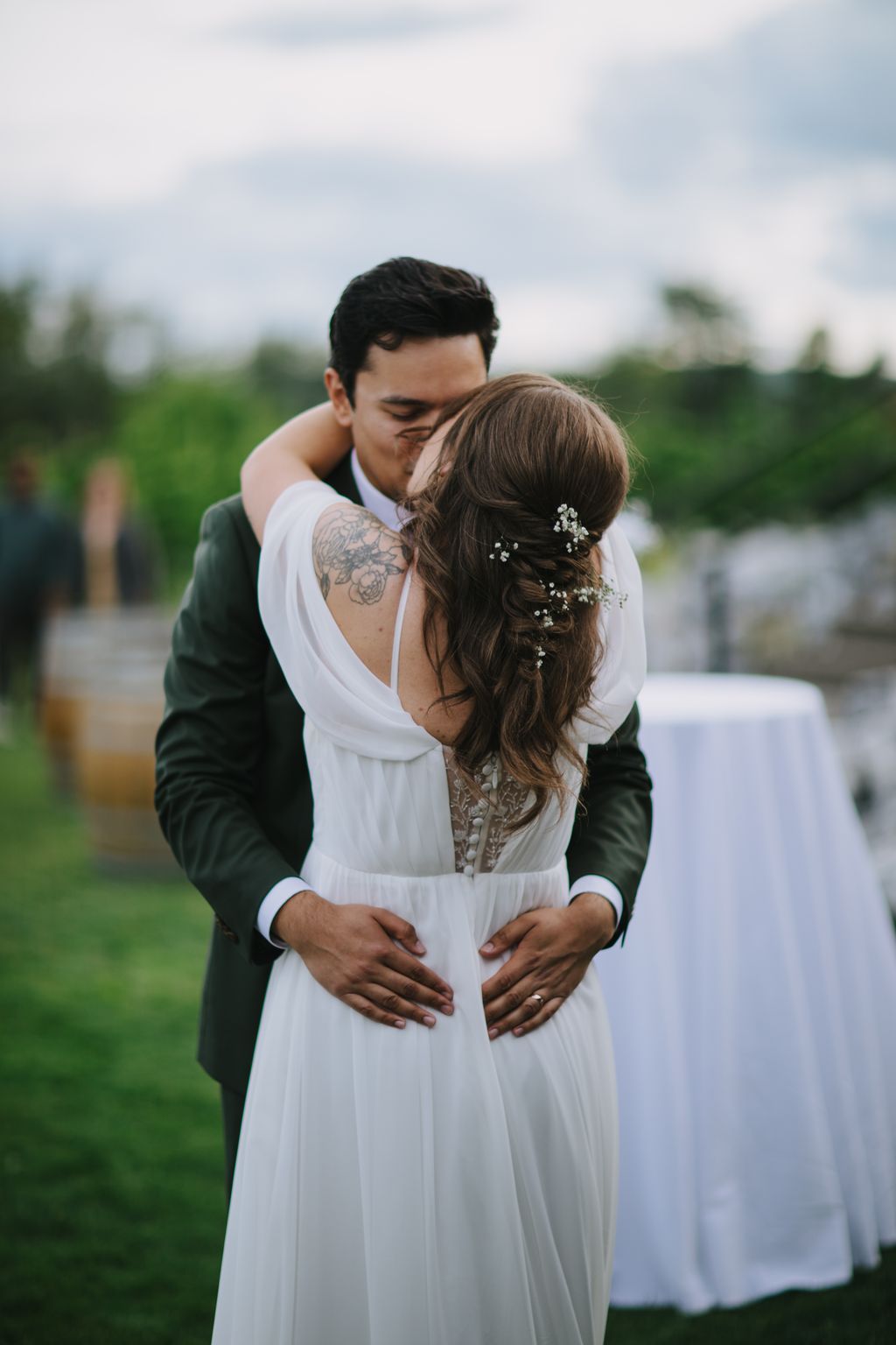 Groom embraces bride from behind in white dress with floral hair pins, outdoor garden setting with soft natural light