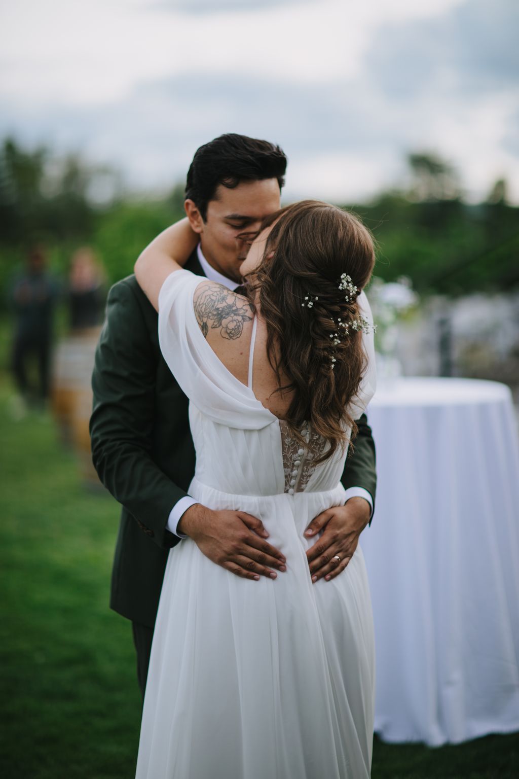 Groom with visible tattoo embraces bride in white dress outdoors during golden hour, green fields and cloudy sky behind them