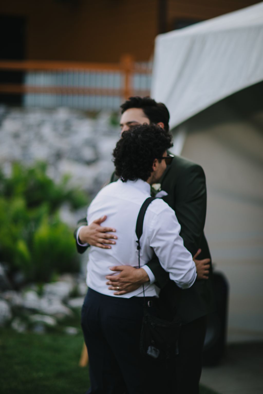Two grooms embrace outdoors, one in a white shirt and one in dark green, with soft natural lighting and blurred architecture behind them