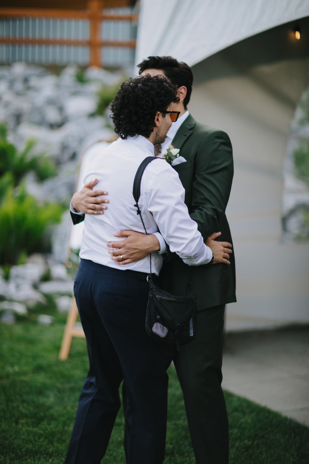 Two grooms embrace outdoors in formal attire, one wearing a green blazer and sunglasses with a boutonniere, in soft natural light