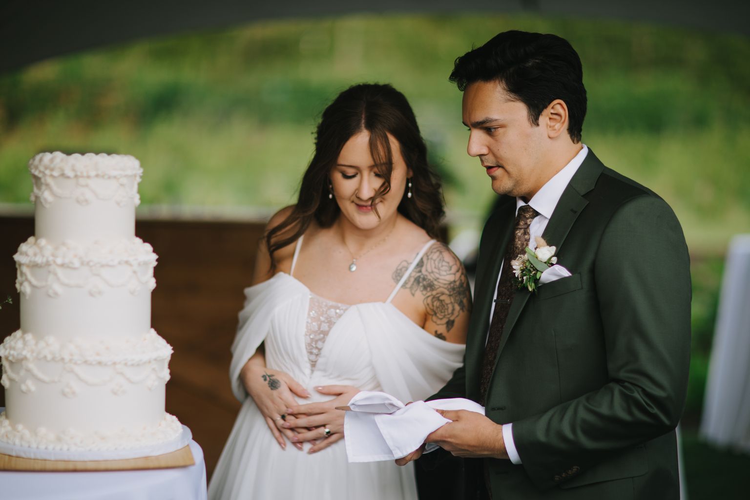 Bride in white dress with shoulder tattoos reads vows while groom in dark suit listens against soft green background