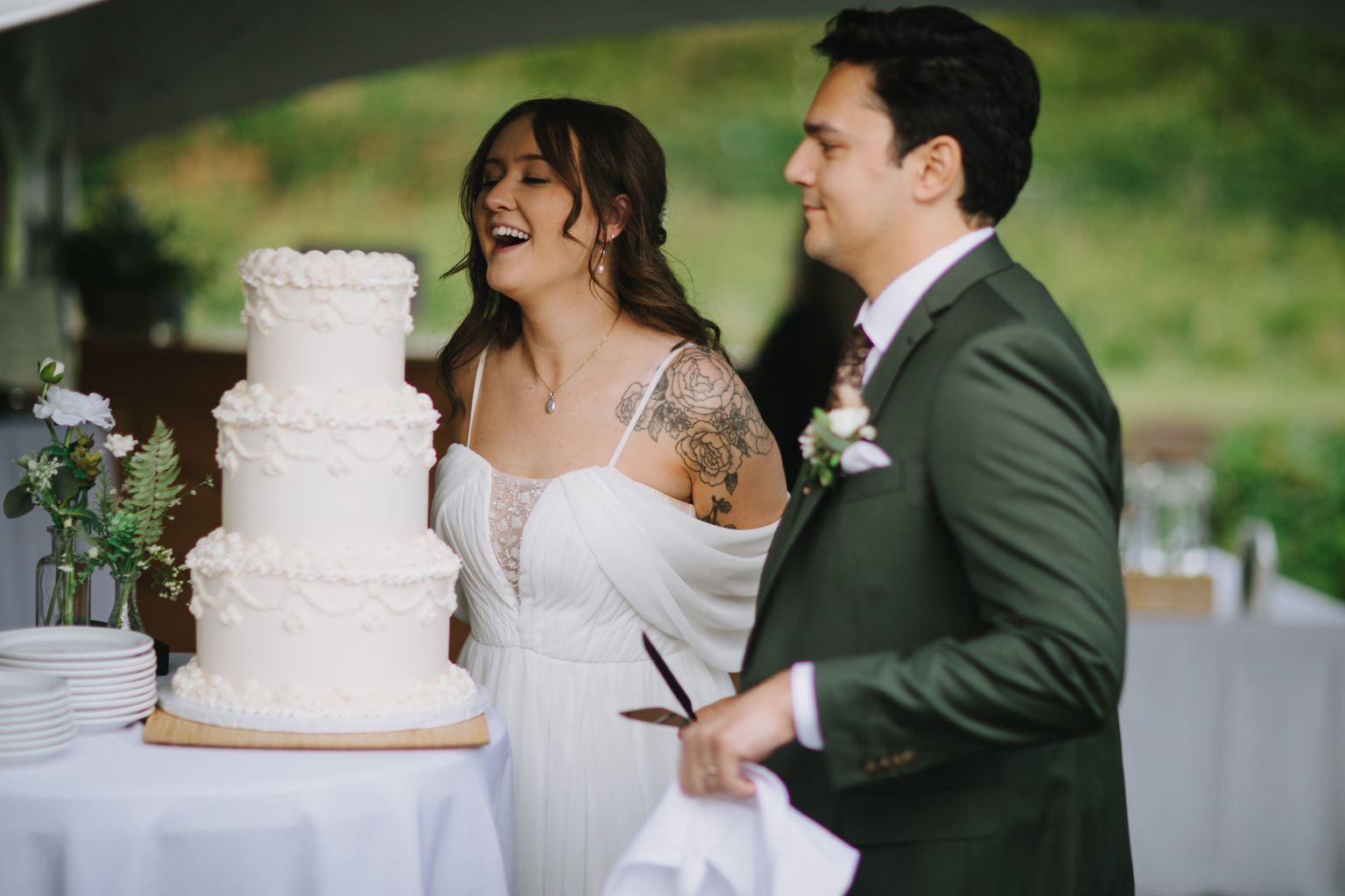 Bride in white dress with shoulder tattoos laughs joyfully while standing with groom in green suit against blurred green background